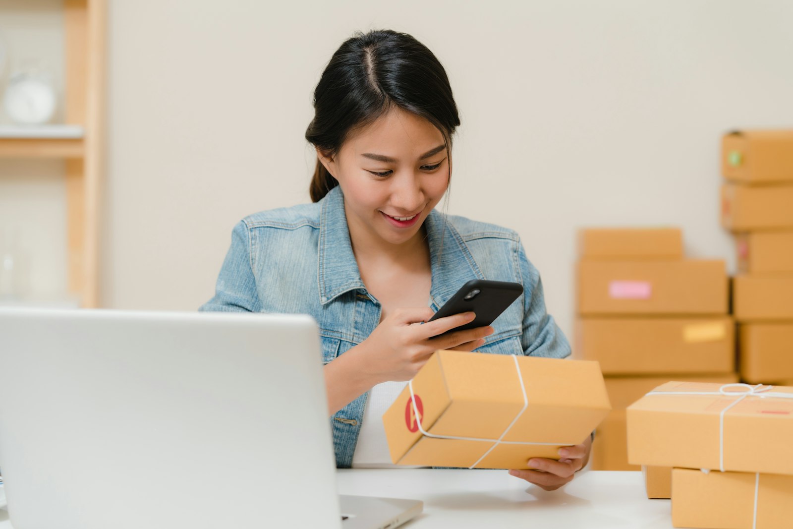 Woman photographing a shipping box with her phone next to a laptop and stacked parcels, the daily routine of a small TikTok Shop seller