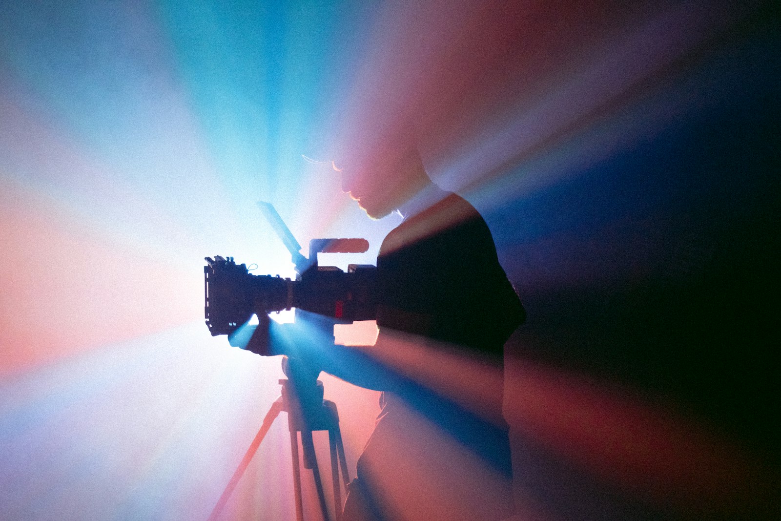 Silhouette of a camera operator filming on a tripod against blue and red studio light, the production cost that sits outside the YouTube media bill